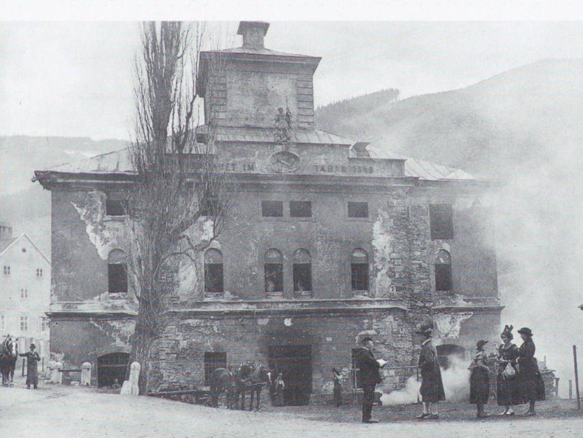 Dieselbe Fassade um 1930: bröckelnder Putz, leere Fensterhöhlen, davor ein Pferdefuhrwerk und Frauen in Tracht